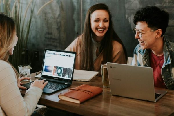 Three woman, smiling, sitting at a desk, working on their laptop