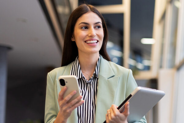 Young Businesswoman Walking In Office Corridor, Holding Laptop And Using Smartphone, Looking Away And Smiling