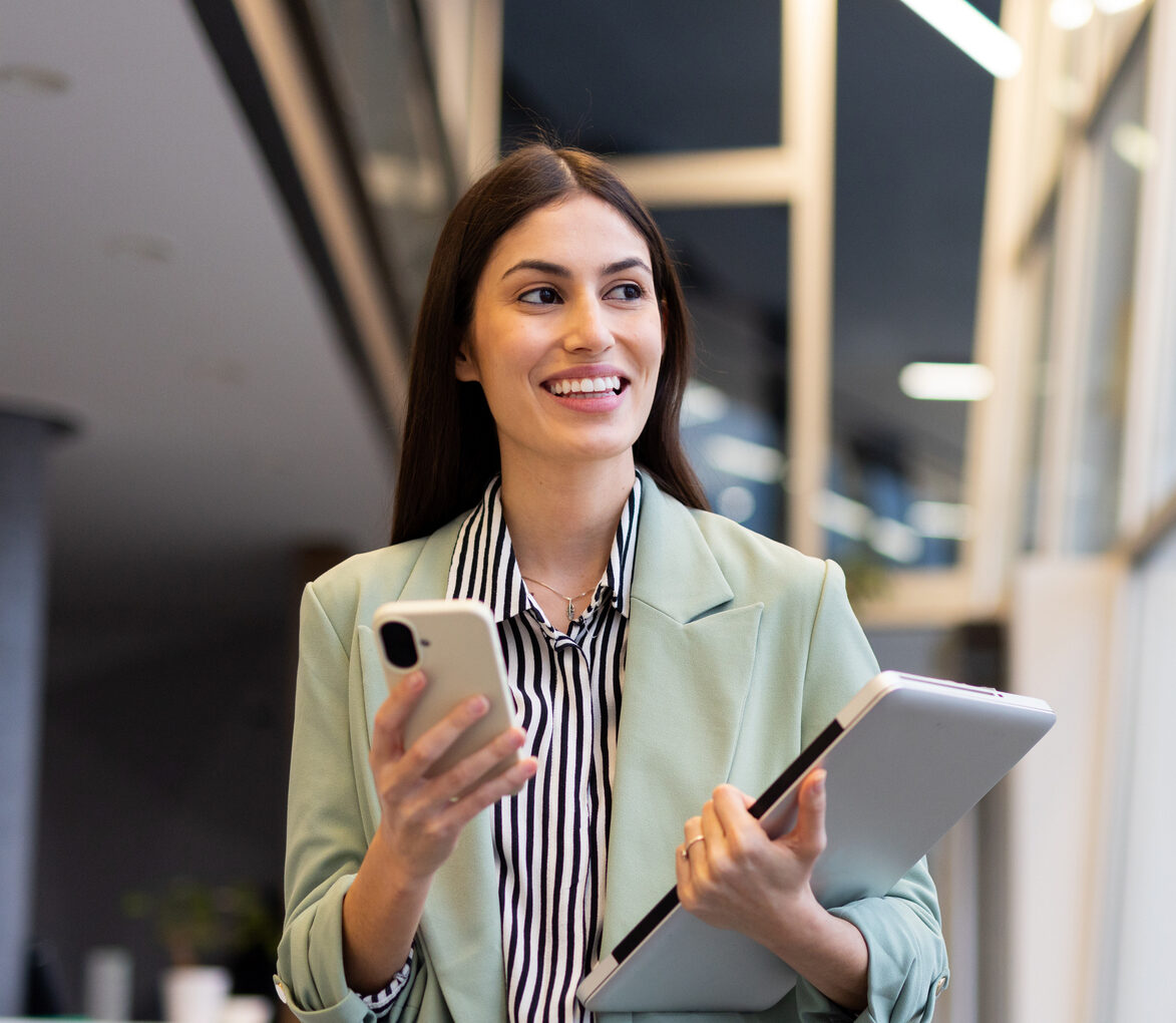 Young Businesswoman Walking In Office Corridor, Holding Laptop And Using Smartphone, Looking Away And Smiling